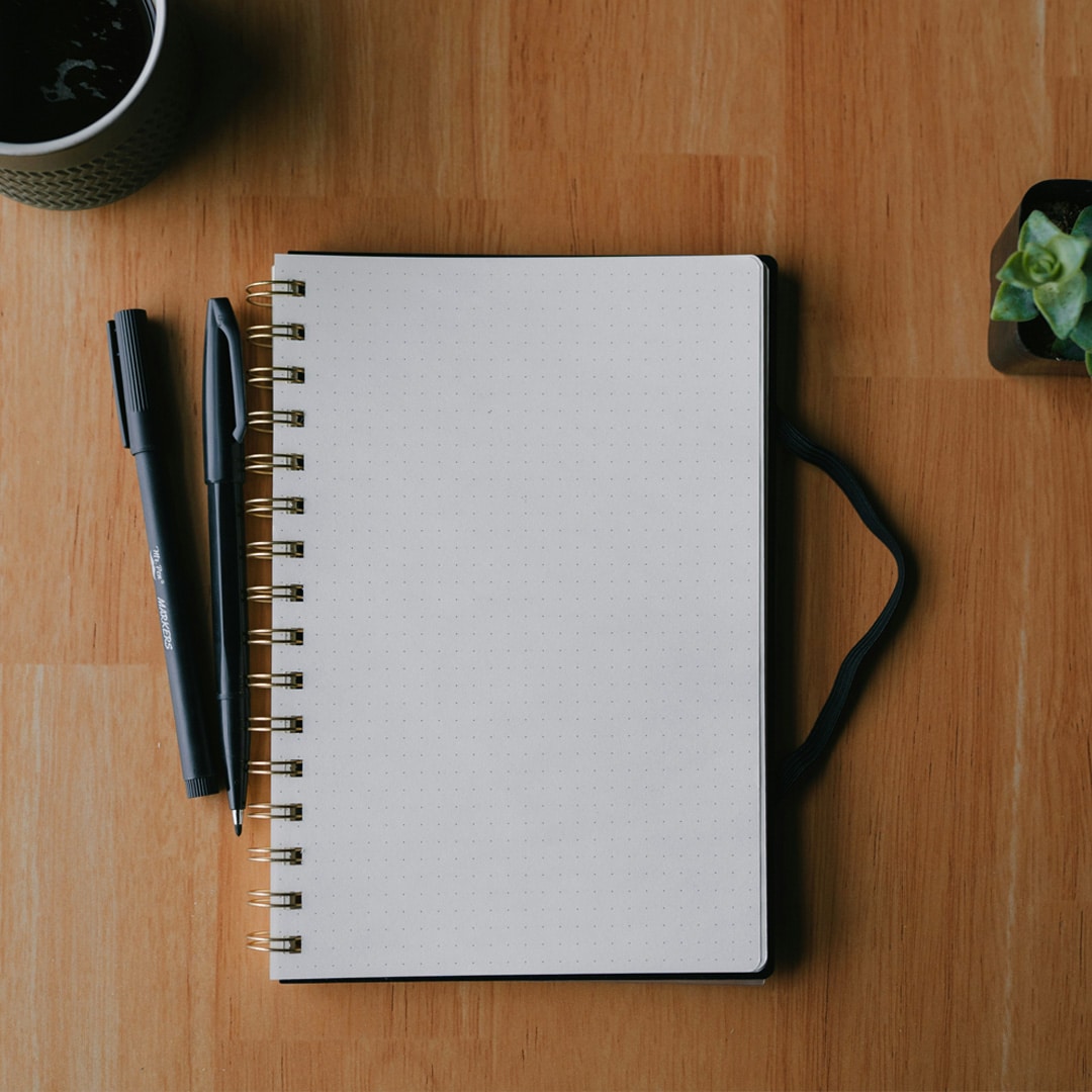 Carnet ouvert avec stylos et tasse de café sur un bureau en bois, illustrant la préparation d’une stratégie marketing digitale.